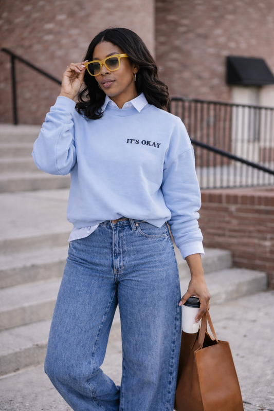 Woman wearing light blue It’s Okay embroidered sweatshirt with jeans and tote bag styled in casual streetwear look for Shop Jayla.